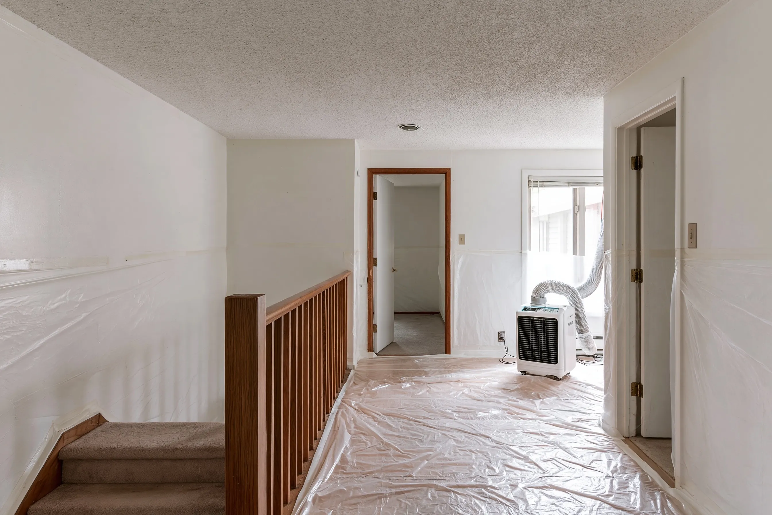 Upper hallway of a split-level with a textured ceiling, plastic on the floor and walls, a filtered air unit vented to a window and the stair rail beside it
